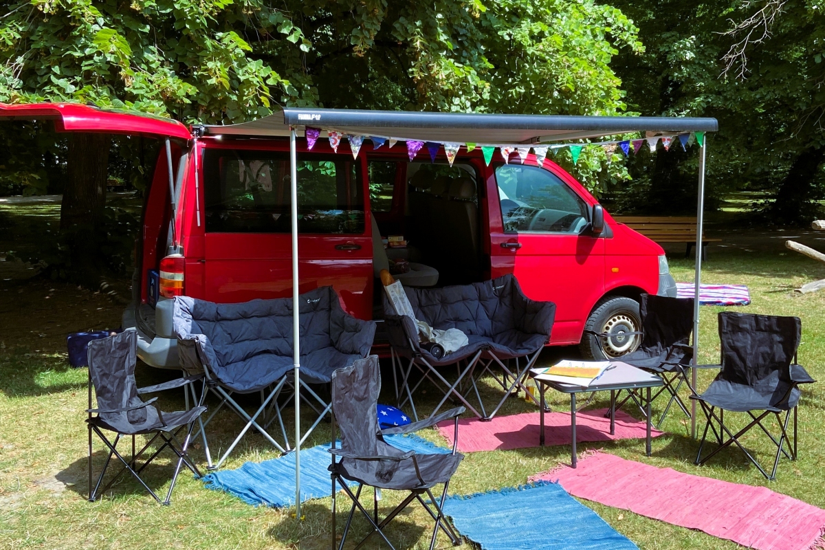 Hier sieht man ein Bild von unserem Streetwork Wohnzimmer. Zu sehen ist unser roter VW Bus mit ausgefahrener Markise, Campingstühle und Sofas im Stadtpark