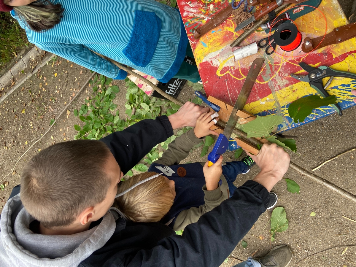 Ein Spielmobilmitarbeiter hält einen Stock, den ein Kind durchsägt. Das Bild ist aus der Perspektive von oben aufgenommen. 