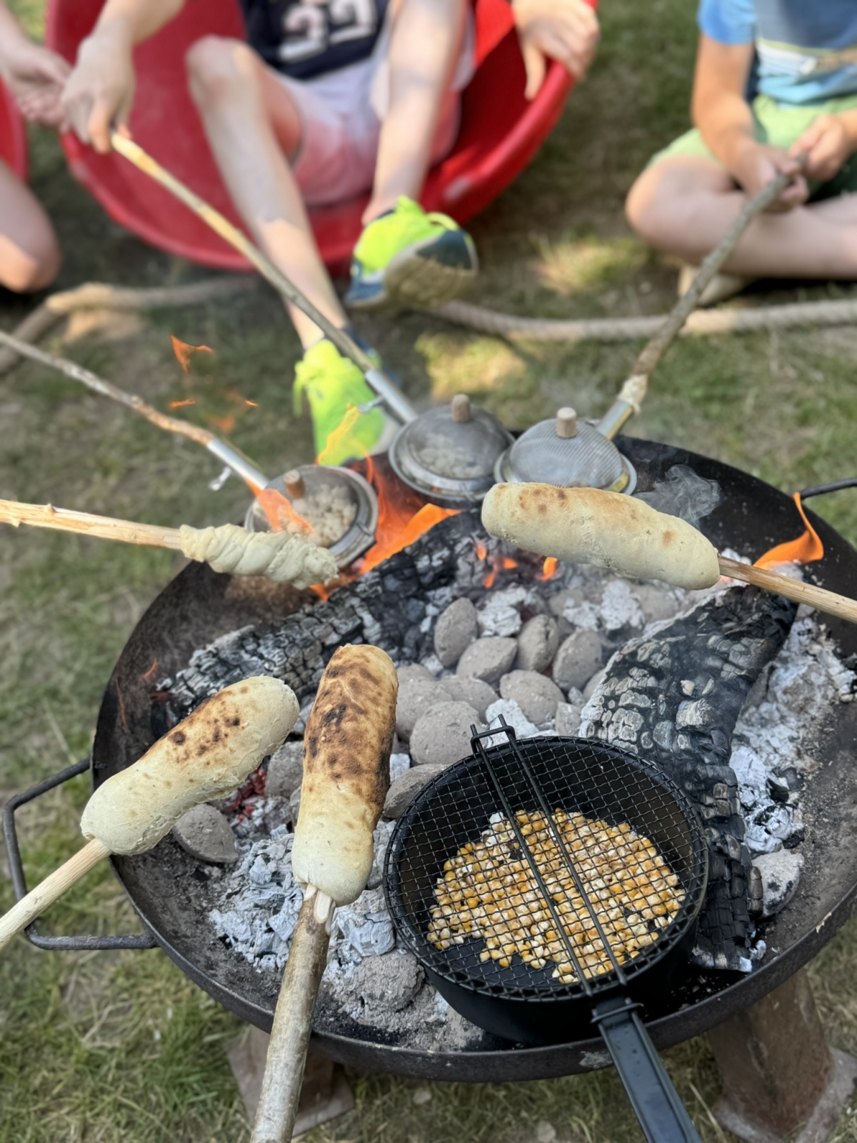 Zu sehen ist das Lagerfeuer über dem Stockbrote gebacken werden und in einer Popcornpfanne Popcorn gemacht wird.