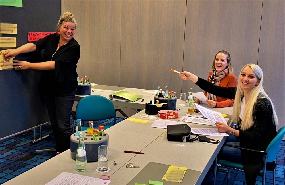 Sabine Heidler pinnt Zettel an eine Pinnwand. Magdalena (Vorsitzende des Vorstands) und eine Mitarbeiterin sitzen gegenüber und lächeln in die Kamera.