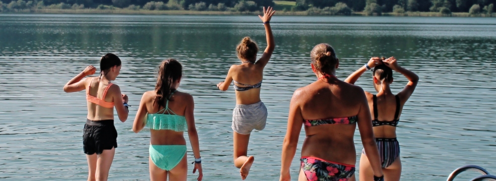 Zu sehen sind eine Gruppe Jugendliche, von hinten in Badebekleidung, die von einer Badeinsel in den See spirngen.