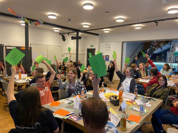 Die Delegierten sitzen an Gruppentischen im Saal des Jugendzentrums Aurex und halten grüne Karten zur Abstimmung hoch.